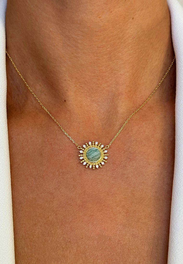 Collier en or avec un pendentif rond en pierre verte entouré de petits cristaux transparents, porté sur une peau bronzée avec des bordures en tissu blanc.