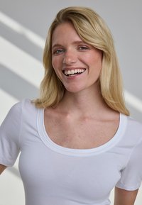 Uśmiechnięta blondynka o niebieskich oczach, ubrana w białą bluzkę z dekoltem w łódkę, stojąca w miękkim, naturalnym świetle z paskami cieni w tle.