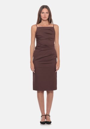 Donna in piedi di fronte, che indossa un vestito marrone aderente senza maniche fino al ginocchio e tacchi neri con cinturino, su uno sfondo semplice.