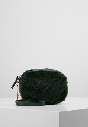 Petit sac à main vert foncé à texture peluche, détail chaîne dorée et bandoulière en cuir, posé sur un socle blanc sur fond gris clair.