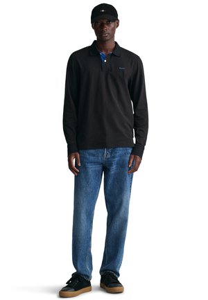 Schwarzes Langarm-Polo mit blauer Kragen, GANT-Logo, kombiniert mit hellblauen Jeans und schwarzen Sneakers mit Gummisohle. Schwarze Kappe getragen.