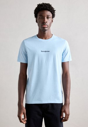 Hellblaues Baumwoll-T-Shirt mit kurzen Ärmeln und Rundhalsausschnitt, auf der Vorderseite mit "Buongiorno" in schwarzer Schrift bedruckt. Kombiniert mit dunklen Hosen.