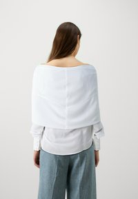 Superposition de tissu blanc et transparent drapée sur les épaules, associée à une chemise blanche ajustée et à un pantalon en denim bleu clair ample.