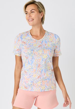 Femme souriante aux cheveux blonds courts portant un T-shirt à motifs floraux multicolores pastel et un short rose clair, debout devant un fond uni.