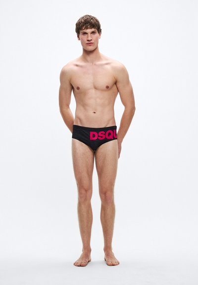 Črne plavalne kratke hlače z teksturiranim vzorcem, na pasu je izrazit roza napis "DSQUARED2". Model je bos in stoji pokončno.