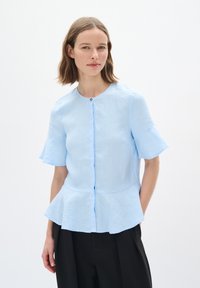 Světle modrá bavlněná blůza s krátkými rozšířenými rukávy, kulatým výstřihem a peplum lemem. Obsahuje zapínání na knoflíky v přední části a jemnou texturu.