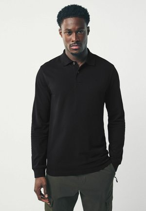 Camisa polo negra de manga larga con cuello y un pequeño logo. Presenta una textura suave y un diseño ajustado, combinada con pantalones verde olivo.