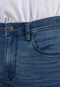 Denim kavbojke z temno modrim pranjem. Imajo kovinsko gumb na pasu, pet žepov in vidno šivanje ob šivih.