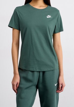 Nainen, joka pukeutunut tummanvihreään Nike-T-paitaan ja vastaaviin collegehousuihin valkoisilla logoilla, seisoo yksinkertaisen vaalean taustan edessä.