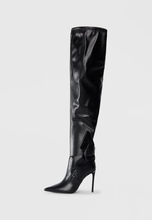 Bota negra de muslo alto hecha de material sintético brillante. Cuenta con una punta afilada, tacón de aguja y un gran lazo decorativo en el lateral.