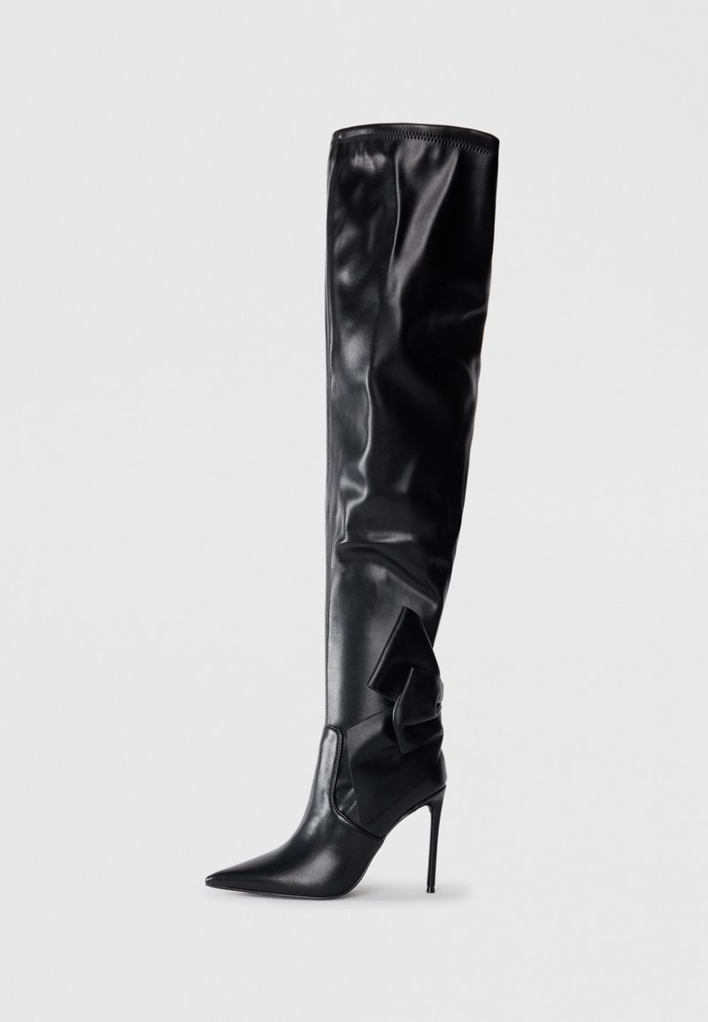 Botte noire haute jusqu'à la cuisse en matériau synthétique brillant. Dotée d'un bout pointu, d'un talon aiguille et d'un grand nœud sur le côté.