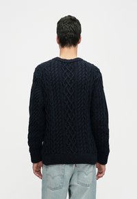 Moški v temno modri puloverju z pletenim vzorcem in svetlih modrih kavbojkah stoji s hrbtom obrnjeno proti kameri na enobarvnem ozadju.