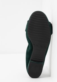 Semelle d'une chaussure en daim vert foncé reposant verticalement contre un mur blanc, montrant une semelle extérieure en caoutchouc noir rainurée et un talon.