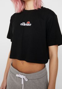 Svart kort t-shirt i bomull. Har en vit broderad "ellesse"-logotyp och en färgglad accent ovanför texten. Enkelt design med korta ärmar.
