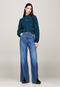 Suéter de punto color verde azulado con acabado texturizado, combinado con vaqueros azules de talle alto y pierna ancha con aberturas en el bajo, y botines negros hasta el tobillo.