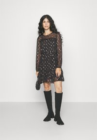 Μαύρο διαφανές φόρεμα με μακριά μανίκια και φουσκωτό στρίφωμα, διακοσμημένο με μικρές, μεταλλικές χρυσές λεπτομέρειες, και συνδυασμένο με μαύρες μπότες μέχρι το γόνατο.