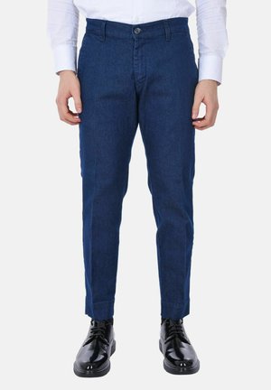 Uomo con pantaloni blu scuro slim fit, camicia bianca con bottoni e scarpe eleganti nere lucide, in piedi con le mani rilassate.