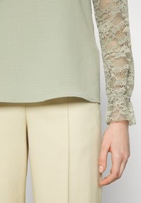 Top verde claro com tecido texturizado e mangas longas de renda transparente, combinado com calças de cor creme.