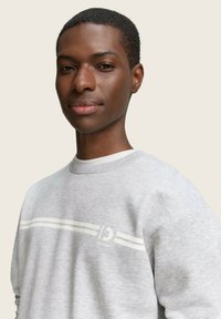 Pilkas džemperis su apvalia kaklo iškirpte, su baltais dryžiais ir logotipu centre. Minkšta tekstūra, laisvas siluetas ir ilgos rankovės.