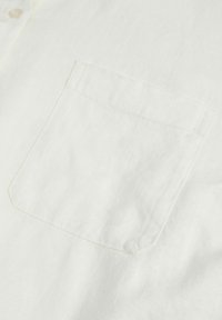 Camicia bianca di lino con taschino sul petto visibile, chiusura a bottoni e una sottile texture, rifinita con cuciture leggere attorno alla tasca.