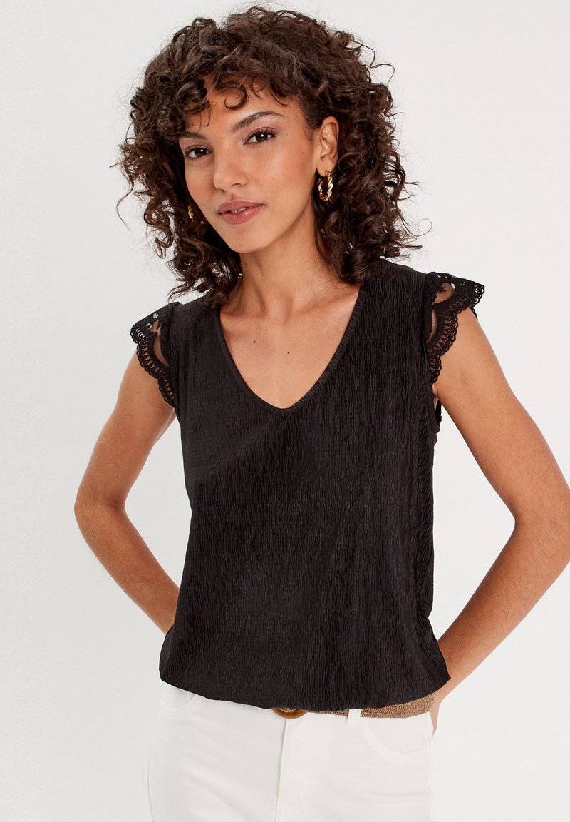 Haut sans manches noir avec un col en V et un tissu texturé. Présente des bordures en dentelle sur les épaules. Porté avec un pantalon blanc et une ceinture marron.