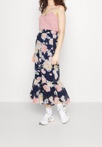 Top sin mangas rosa combinado con una falda maxi floral azul marino que presenta grandes flores rosas y blancas y un dobladillo con volantes, vestida con zapatillas blancas.