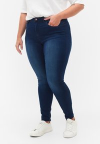 Jeans ajustados de mezclilla azul oscuro, con una cintura alta, bolsillos delanteros y cierre de botón. Combinados con zapatillas blancas.