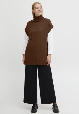 Braune Strick-Tunika mit hohem Kragen, kurzen Ärmeln und lockerer Passform, kombiniert mit weiten schwarzen Hosen und schwarzen Schuhen. Glatte Textur.