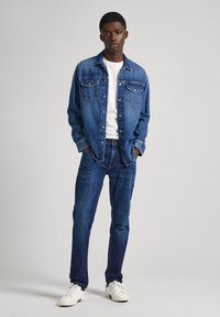 Denim jakna u srednje plavoj boji s dva džepa na prsima, u kombinaciji s bijelom majicom i tamnoplavim trapericama. Bijele tenisice upotpunjuju cjelokupni izgled.
