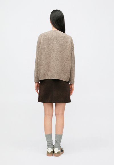 Femme aux longs cheveux noirs portant un pull beige, une jupe marron foncé, des chaussettes grises et des baskets beiges, debout de dos sur fond blanc.