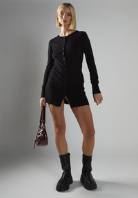 Kobieta z blond bobem w czarnej mini sukience na guziki, trzymająca ciemnoczerwoną torebkę i nosząca czarne masywne botki, stojąca na szarym tle.