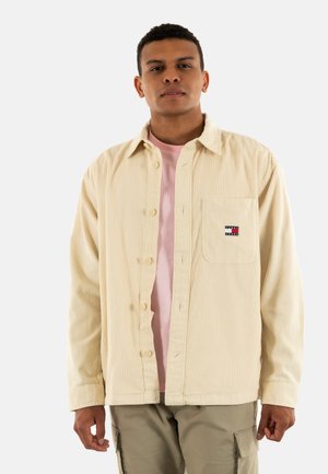 Camisa de botonadura de pana color beige claro con un bolsillo en el pecho, con un pequeño parche de logo en rojo, blanco y azul. Se lleva sobre una camiseta rosa.
