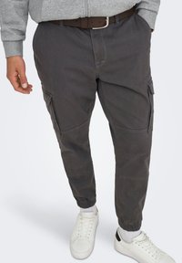 Pantalones cargo gris oscuro con puños elásticos, bolsillos laterales y costuras reforzadas. Combinados con zapatillas blancas y una sudadera con capucha gris con cremallera.
