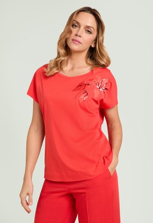 Femme portant un haut rouge à manches courtes avec un détail floral en sequins près de l'épaule et un pantalon rouge assorti, debout avec une main dans la poche.