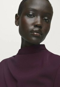 Primo piano di una persona con pelle scura, capelli corti e labbra lucide che indossa un top a collo alto di un viola scuro su uno sfondo semplice.