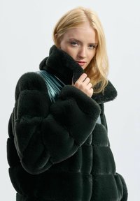 Blondynka poprawiająca kołnierz puchowej, ciemnozielonej kurtki o pikowanej strukturze, patrząc w kierunku kamery.