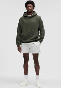 Un hoodie vert olive avec une poche avant, associé à un short gris clair et des baskets blanches. Le mannequin a les cheveux courts et bouclés et se tient debout.