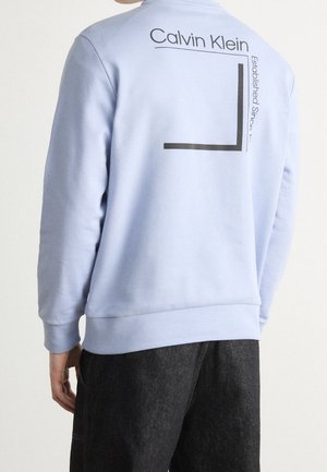 Person trägt ein hellblaues Sweatshirt mit schwarzem Text und Grafik "Calvin Klein Established Since 19--" auf dem oberen Rücken, kombiniert mit dunkler Hose.