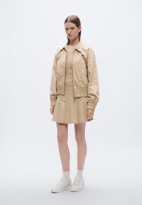 Beige bomberjack met geribbelde boorden, gecombineerd met een plissé beige rok. Model draagt witte sneakers en sokken. Soepele, gestructureerde stof.