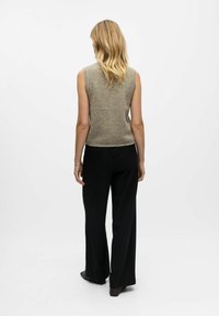 Sieviete ar blondiem matiem, ģērbusies bez piedurknēm bēšā džemperī un melnos svītrainos platās biksēs, stāv vērsta pret baltu fonu.