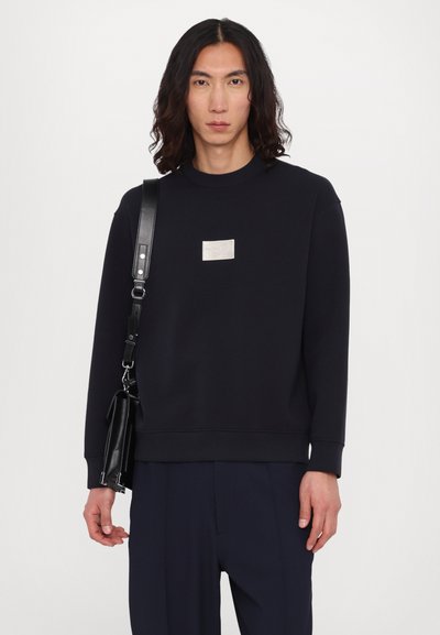 Sweatshirt bleu marine en tissu doux, avec un col rond et un patch logo argenté sur la poitrine. Le modèle tient un sac noir.