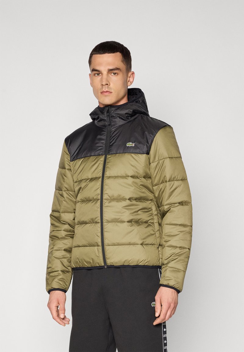 Giacca puffer verde oliva con cappuccio e spalle nere, caratterizzata da motivi a righe orizzontali, zip frontale e logo discreto sul petto.