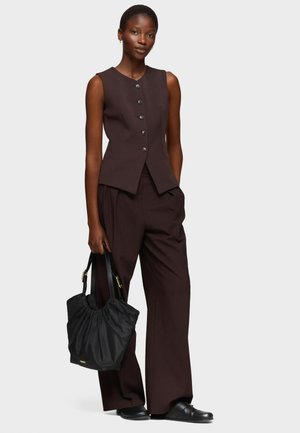 Kobieta ubrana w brązową bezrękawnik i pasujące szerokie spodnie, trzymająca czarną torebkę, stojąca na tle jasnego, jednolitego tła.