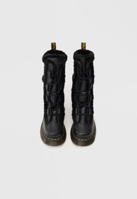 Botas negras con cordones, con un exterior de cuero texturizado, forro de piel de pelaje negro suave, punta redondeada y detalle de costura amarilla en la suela.