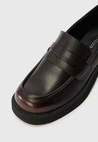 Mocassino in pelle marrone scuro con finitura liscia, caratterizzato da una dettaglio con fascia firmata e suola in gomma nera per una maggiore aderenza.