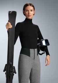 Čierny pletený rolák a sivé textúrované lyžiarske nohavice so zeleným pásom. Drží čierne lyže a čiernu lyžiarsku prilbu s reflexným zorníkom.