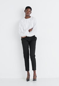 Blusa bianca texturizzata con design ondulato, abbinata a pantaloni neri su misura. La modella indossa orecchini a cerchio in argento e scarpe nere con punta.