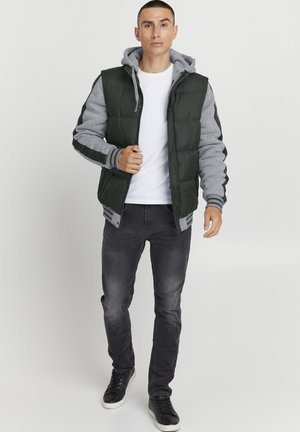 Padded jacket z ciemnozielonym zewnętrzem i szarymi rękawami. Posiada szary kaptur, zamek błyskawiczny z przodu oraz prążkowane ściągacze. Noszony z szarymi dżinsami i trampkami.