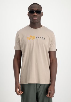 Bežna bombažna majica s kratkimi rokavi, ki na prsih prikazuje črno in rumeno "Alpha Industries" logotip z geometrskim oblikovanjem. Ima okroglo izrezano ovratnik.