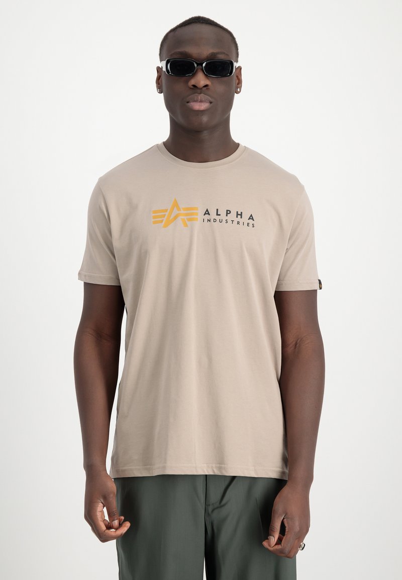 Camiseta de algodón beige con un logotipo negro y amarillo de "Alpha Industries" con un diseño geométrico en el pecho. Mangas cortas y cuello redondo.